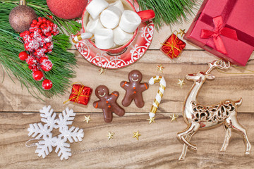 Christmas time. A mug with a snowman, deer, marshmallow and gingerbread cookies on a wooden table is decorated with Christmas decorations, fir branches, toys with a gift.