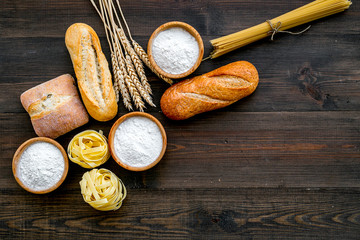 Farinaceous food. Fresh bread and raw pasta near flour in bowl and wheat ears on dark wooden background top view space for text