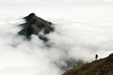An adventurous male traveler looks out over a mountain peak rising above a thick layer of clouds in...