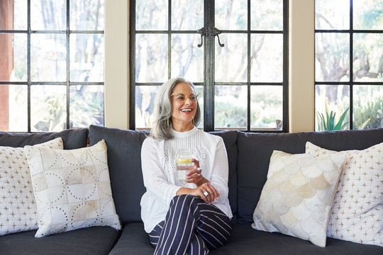 Portrait of mature woman with grey hair laughing in her living room at home holding a glass of water