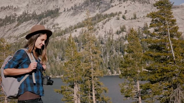 Young beautiful tourist girl with backpack and camera smiling at camera while hiking at Yosemite park lake slow motion.