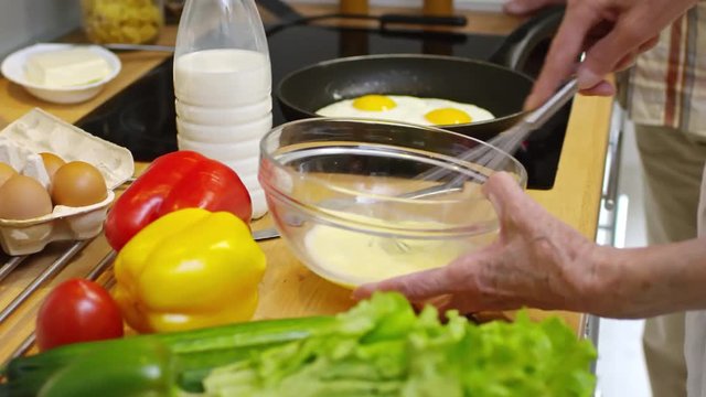 Close up of hands of old person mixing eggs in big plate for omelet and somebody frying scrambled eggs on oven