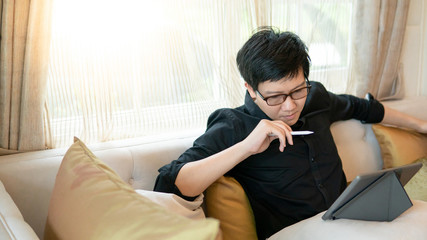 Young Asian business man with eyeglasses using digital tablet while sitting on sofa or couch in living room. Home living lifestyle with modern electronic gadget concept