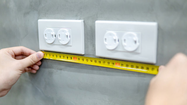 Male hand electrician using tape measure for measuring dimension of electrical outlet with safety cover on the wall. Electrical fixture installation and building construction concepts