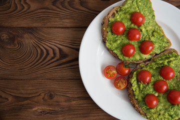 Healthy avocado sandwich on dark rye toast bread made with fresh avocado paste, cherry tomatoes on brown wooden background