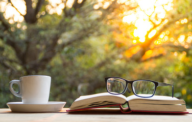 A cup of coffee and glasses on book on the wooden table with morning light and blurred nature...