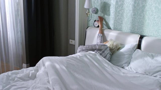 Tracking right of old woman lying on her back in bed, discontentedly turning off alarm clock and continuing sleep in the morning