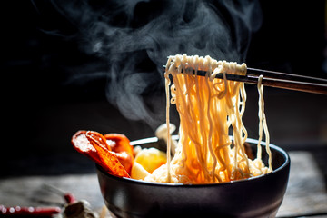 Hand uses chopsticks to pickup tasty noodles with steam and smoke in bowl on wooden background, selective focus. Asian meal on a table, junk food concept