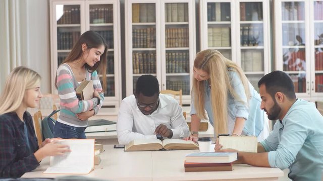 Mixed-race group of university students with a focus on blonde caucasian woman doing linguistic researches, turning over the pages of big old reference textbook at library.