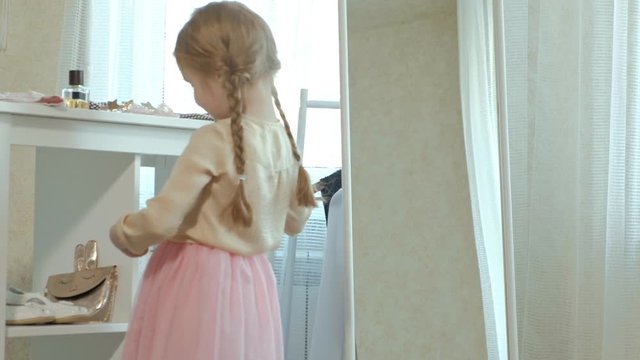 cheerful little girl with pigtails in a pink skirt paints her lips with lipstick in front of the mirror, her mother wardrobe
