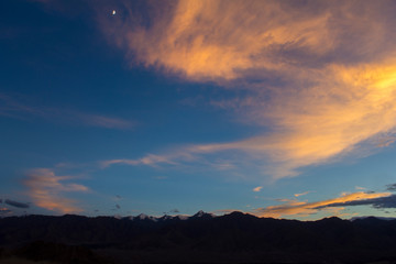 bright clouds in the dark sky and the moon over the silhouettes of mountains with snowy peaks