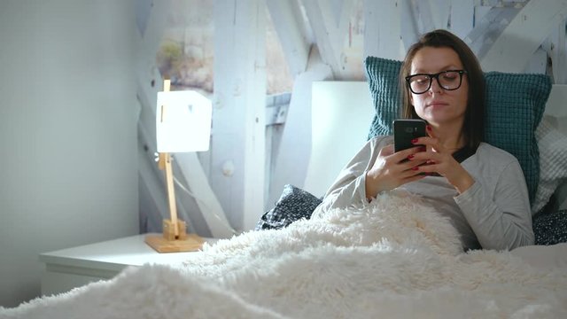 Woman in glasses lies in a bed and uses a smartphone before bedtime