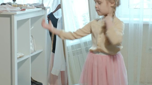 cheerful little girl with pigtails in a pink skirt tries on mother's shoes on heels and dances in front of a mirror with a bag, mother's wardrobe