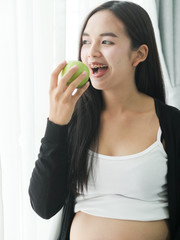 Happy Asian woman eating green apple near window at home, lifestyle concept.