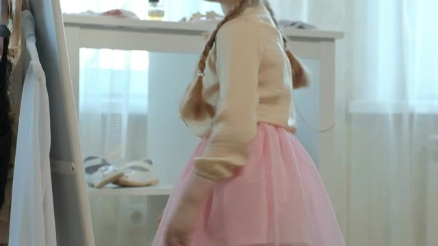 cheerful little girl with pigtails in a pink skirt tries on mother's shoes on heels and dances in front of a mirror, mother's wardrobe