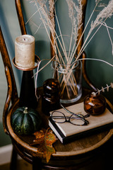 A wooden table decorated with an autumn theme featuring an aromatic candle, glasses, dried flowers, an oak leaf, and a green pumpkin