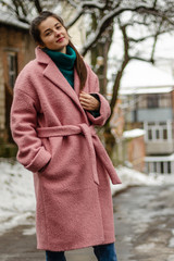 beautiful brunette in a warm pink coat on a cold, gloomy street poses with a smile for a photo, an...
