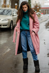 beautiful brunette in a warm pink coat on a cold, gloomy street poses with a smile for a photo, an...