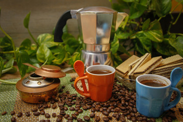 Orange and blue cups of coffee, moka pot coffee maker, old alcohol stove and cookies. Wooden table with bamboo mat and coffee beans spilled on the table.