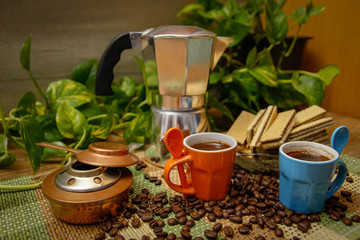 Orange and blue cups of coffee, moka pot coffee maker, old alcohol stove and cookies. Wooden table with bamboo mat and coffee beans spilled on the table.
