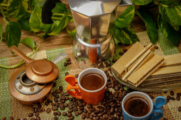 Orange and blue cups of coffee, moka pot coffee maker, old alcohol stove and cookies. Wooden table with bamboo mat and coffee beans spilled on the table.