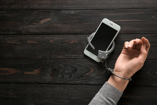 Female hand in handcuffs with mobile phone on wooden background. Concept of addiction