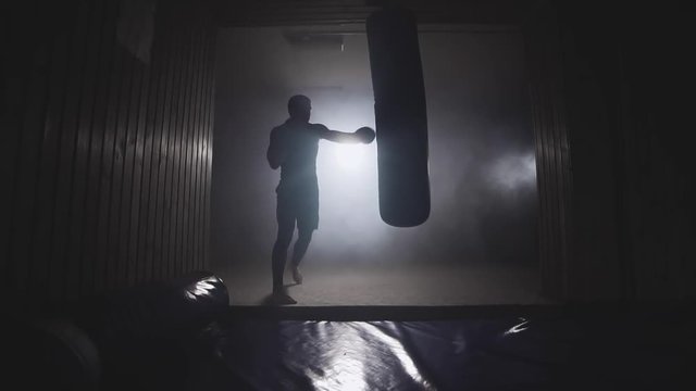 Alone boxer hits punching bag in dark gym. Young man training indoors. Strong athlete in gym. Sport concept. Medium shot. Sportsman boxing in smoky studio