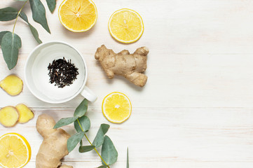 Fresh ginger root, cup tea with brewing inside, lemon, eucalyptus leaves on white wooden background. Flat lay, top view, copy space. Minimalistic style, seasoning, spice, ingredient for tea.