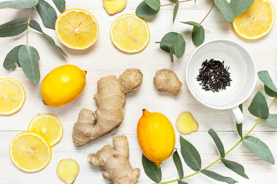 Fresh ginger root, cup tea with brewing inside, lemon, eucalyptus leaves on white wooden background. Flat lay, top view, copy space. Minimalistic style, seasoning, spice, ingredient for tea.