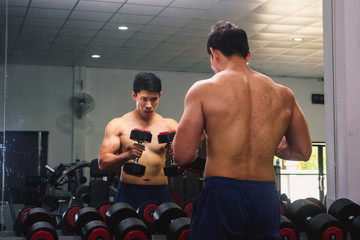An Asian man lifts a dumbbell in front of a mirror in a gym. Exercise in gym concept
