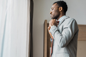 side view of african american businessman adjusting red tie and looking at window