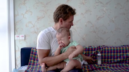 Father starts to feed baby milk from bottle. Reversed gender role with dad feeding infant baby formula. Mixed blood infant sits on Caucasian father's lap. Baby being fed by parent.
