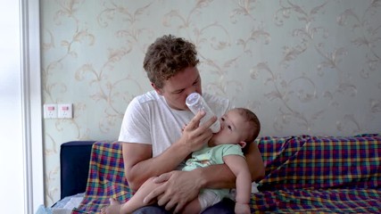 Father finish feeding baby milk from bottle. Reversed gender role with dad feeding infant baby formula. Mixed blood infant sits on Caucasian father's lap. Baby being fed by parent.