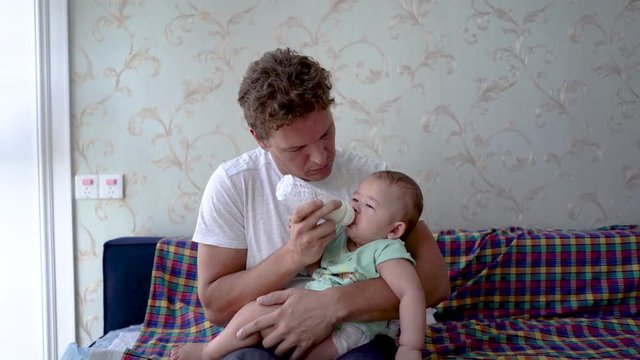 Father feeds baby milk from bottle. Reversed gender role with dad feeding infant baby formula. Mixed blood infant sits on Caucasian father's lap. Baby being fed by parent.