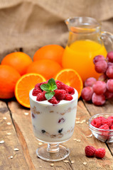 Greek yoghurt with oatmeal and fresh raspberries in a glass, mint leaves, orange juice, grapes in background - vertical photo