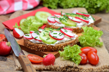 Close-up of grain bread with spread, fresh cucumber, radish, tomatoes and knife on a wooden cutting board - concept of healthy fitness breakfast or snack