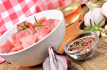 Raw pork meal diced in a bowl, garlic, half of onion, pepper, jug with oil and checkered red tablecloth in the background