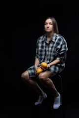 A girl in a shirt is sitting on a chair holding oranges in her hands on a black background.