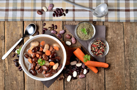 View From Above Of Bowl Of Bean Soup With Large Beans On Cutting Board, Carrots, Parsley, Marjoram, Spoon, Towel And Ladle In Background