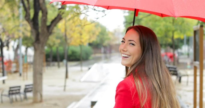 Back view portrait of a happy woman walking smiling looking at camera holding a red umbrella under the rain