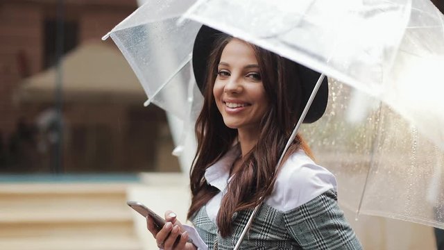 Beautiful young business woman using smartphone on the street in rainy weather, smiling, holding umbrella. She looking into the camera. Communication concept