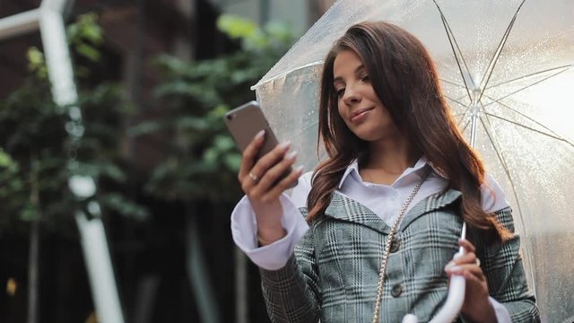 Beautiful young business woman using smartphone on the street in rainy weather, smiling, holding umbrella, communication concept