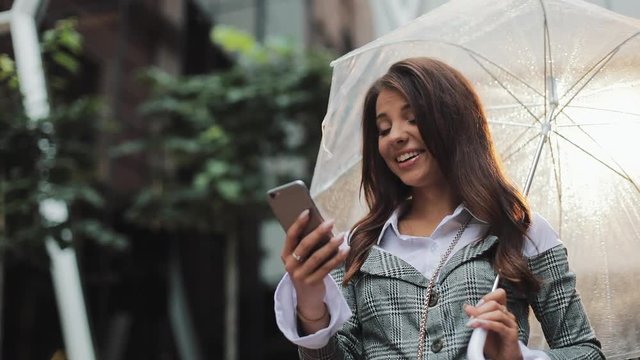 Beautiful young business woman using smartphone on the street in rainy weather, smiling, holding umbrella, communication concept