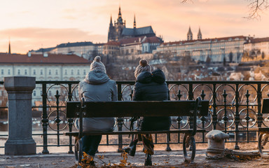 Two women sit on old wooden bench. An astonishing view of the colorful Prague gothic Castle over...