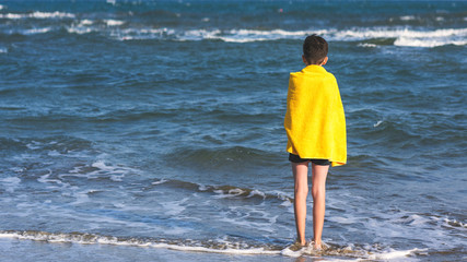 Fototapeta na wymiar Back view on the boy in the yellow towel standing on seashore on the sea backgroung