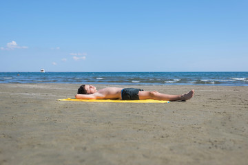 Fototapeta na wymiar Teen boy lies on yellow towel and sunbathes on the beach on the sea and sky background. Concept