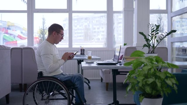 crippled restaurant owner in wheel chair wearing glasses is drinking coffee at a table uses a cellphone and working on a laptop indoors