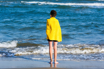 Fototapeta na wymiar Back view on the boy in the yellow towel standing on seashore. Travel Concept.