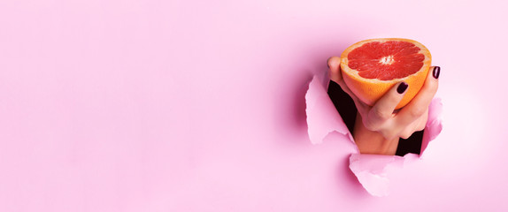 Female hand holding half of grapefruit through torn pink paper background. Fresh orange juice. Vegan, vegetarian concept. Banner with copy space