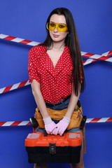 Fototapeta na wymiar A beautiful brunette in a red shirt and yellow glasses is holding a special suitcase for tools on a blue background. Construction concept